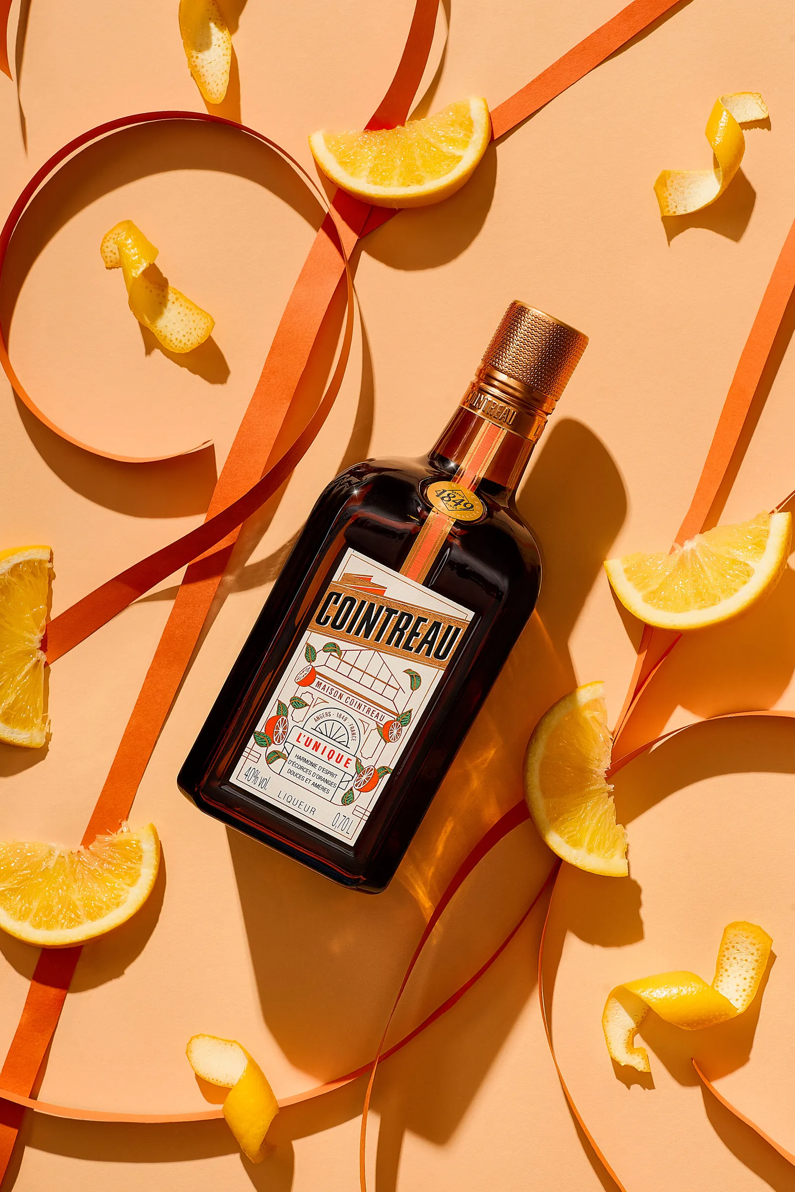 Photo produit : bouteille de liqueur à l’orange Cointreau sur fond pêche, entourée de quartiers d’orange et de rubans de papier