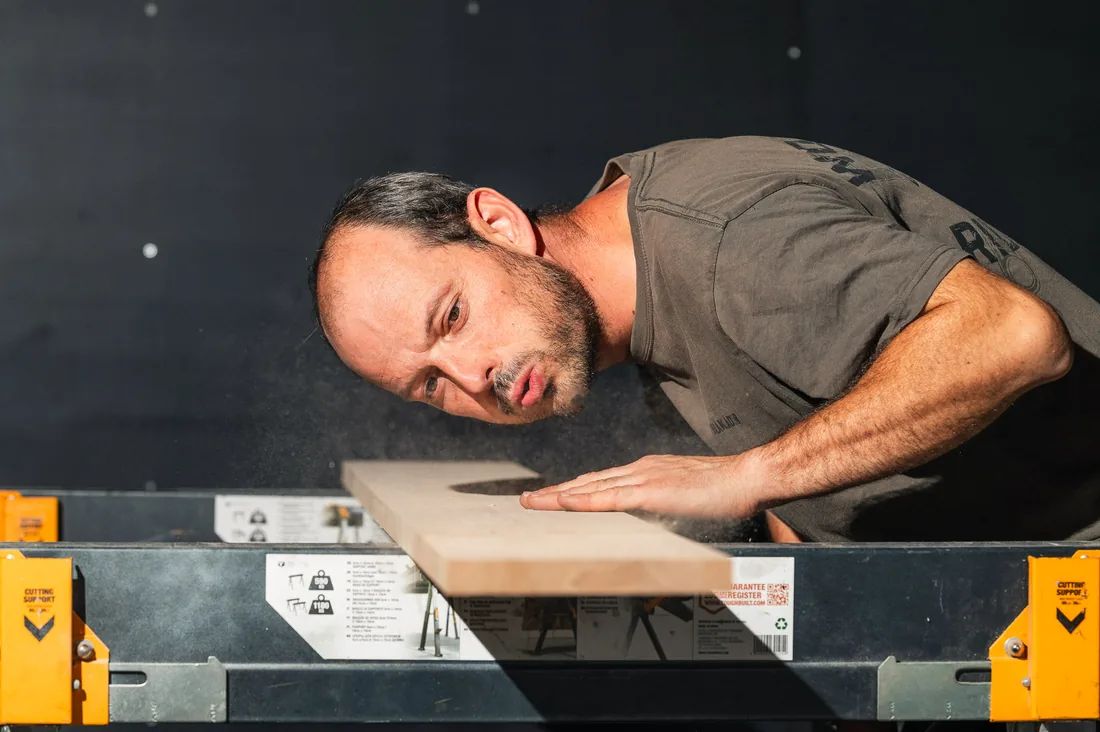 Menuisier soufflant la sciure sur une planche de bois posée sur des tréteaux, en atelier