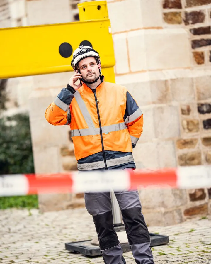 Chef de chantier au téléphone devant un bâtiment en pierre, en tenue haute visibilité